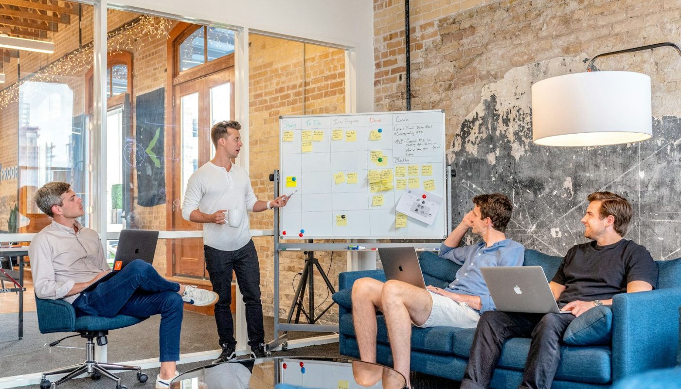 drei Männer sitzen an Laptops und beobachten den Mann neben dem Whiteboard