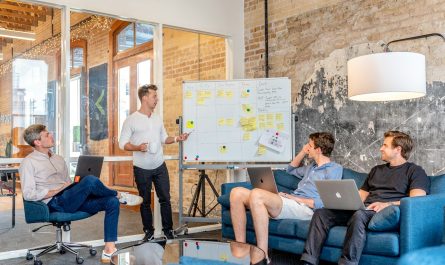 drei Männer sitzen an Laptops und beobachten den Mann neben dem Whiteboard